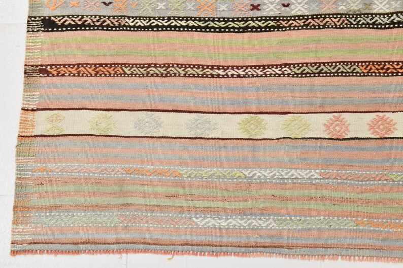 May include: A close-up of a woven rug with a striped pattern in shades of pink, green, brown, and white. The rug has a fringe border.