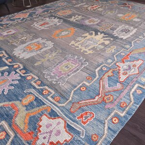May include: A large, rectangular rug with a blue and grey central field and a blue border. The rug features a repeating pattern of stylized floral and geometric designs in orange, purple, and cream colours. The rug is made of a woven material.