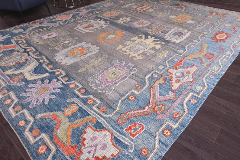 May include: A large, rectangular rug with a blue and grey central field and a blue border. The rug features a repeating pattern of stylized floral and geometric designs in orange, purple, and cream colours. The rug is made of a woven material.