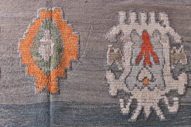 May include: Close-up of a woven textile featuring two decorative patterns. One is an orange and green diamond shape, the other is a white and grey floral-like design with an orange accent. The background is grey and blue.