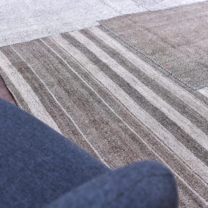 May include: A patchwork rug with a mix of neutral tones, including gray, beige, and brown. The rug features a striped pattern in the center and various rectangular patches. A section of a blue armchair is visible in the foreground.