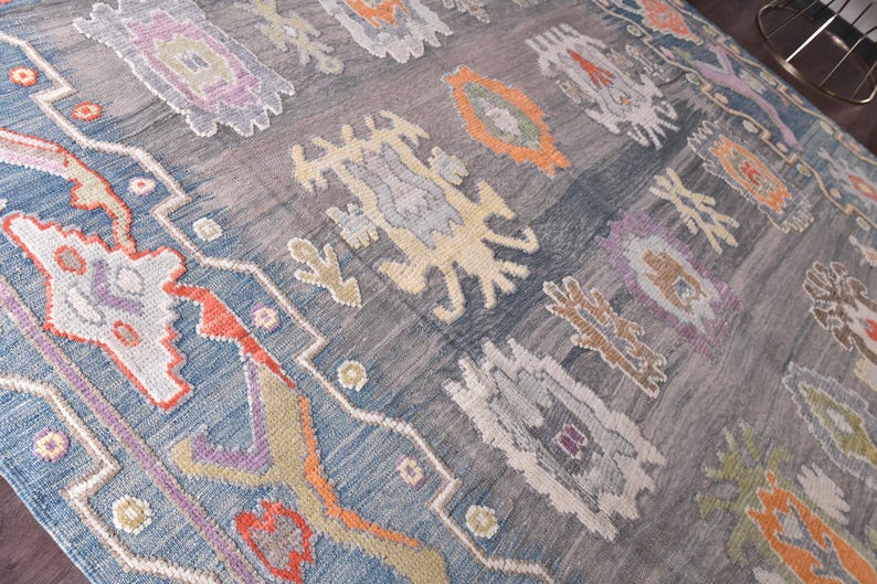 May include: A large, rectangular rug with a faded, distressed look. The rug has a muted colour palette of blues, greys, and accents of orange, purple, and cream. The design includes geometric and floral patterns, creating a vintage aesthetic.