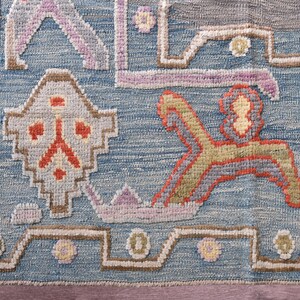 May include: Close-up of a blue, grey, and beige patterned rug. The rug features geometric shapes and floral motifs in shades of orange, yellow, and purple. The rug has a textured appearance.