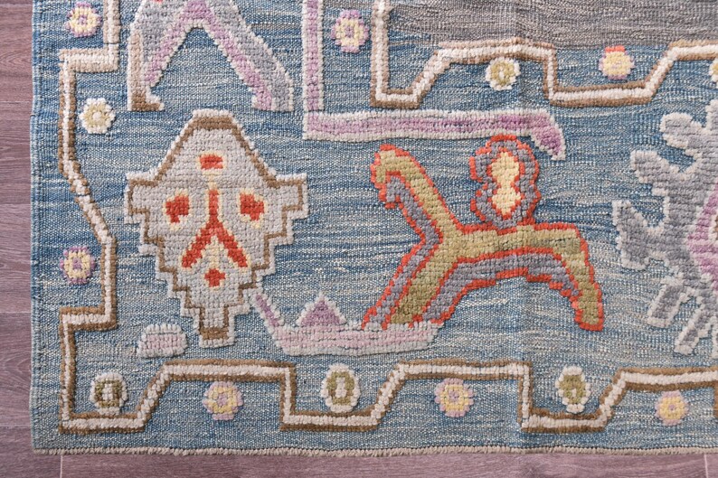 May include: Close-up of a blue, grey, and beige patterned rug. The rug features geometric shapes and floral motifs in shades of orange, yellow, and purple. The rug has a textured appearance.