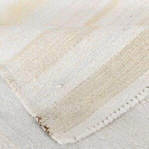 May include: Close-up of a folded, off-white rug with beige stripes. The rug has a textured weave and a decorative, stitched edge. The neutral colour palette and simple design suggest a minimalist aesthetic.
