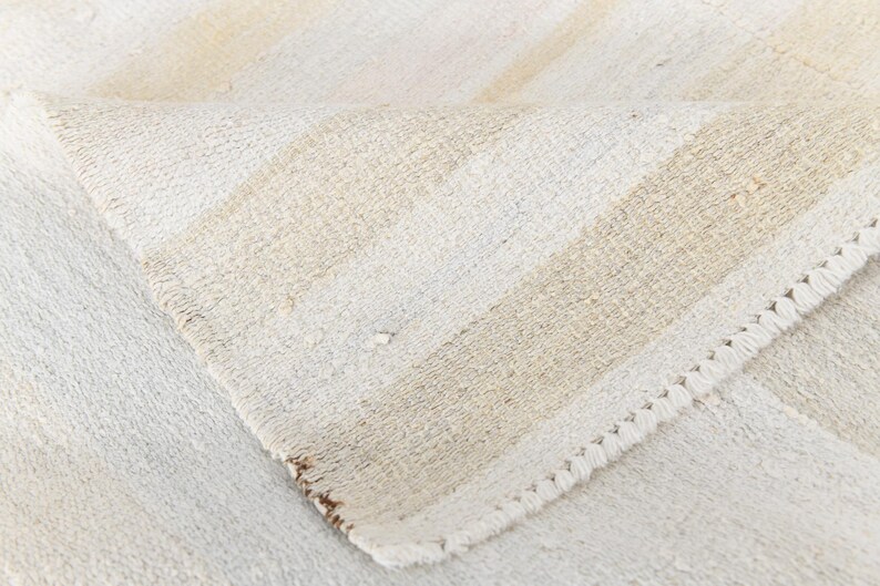 May include: Close-up of a folded, off-white rug with beige stripes. The rug has a textured weave and a decorative, stitched edge. The neutral colour palette and simple design suggest a minimalist aesthetic.