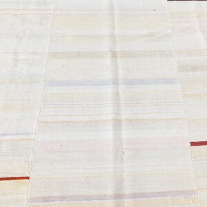 May include: A close-up of a cream-coloured rug with horizontal stripes in various muted colours. The rug has a patchwork design with visible seams. Thin red stripes accent the design.