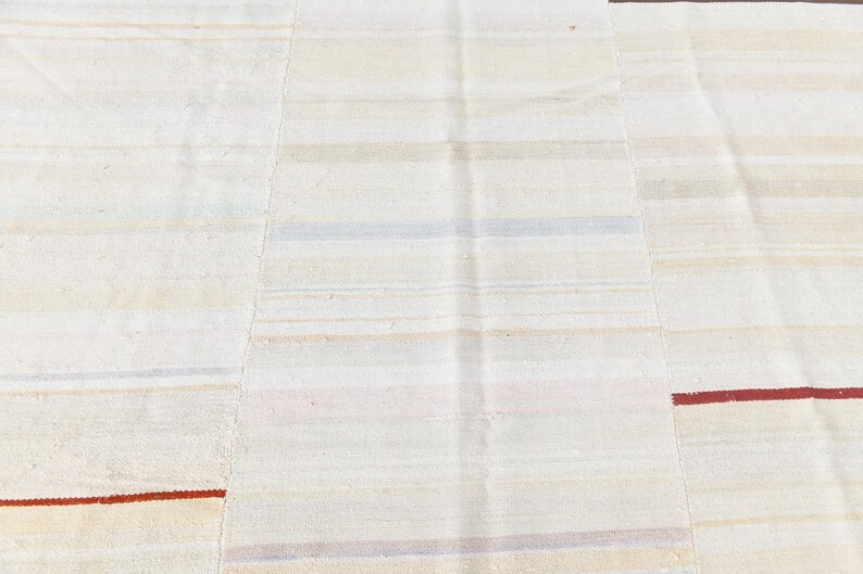May include: A close-up of a cream-coloured rug with horizontal stripes in various muted colours. The rug has a patchwork design with visible seams. Thin red stripes accent the design.