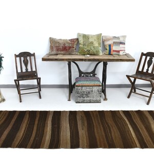 May include: A brown striped rug with fringe on the ends. The rug is in front of a wooden table with three pillows on it. There are two wooden chairs and a small stool with a colorful striped cushion on it. A painting of a woman in a yellow and green dress is on the wall.