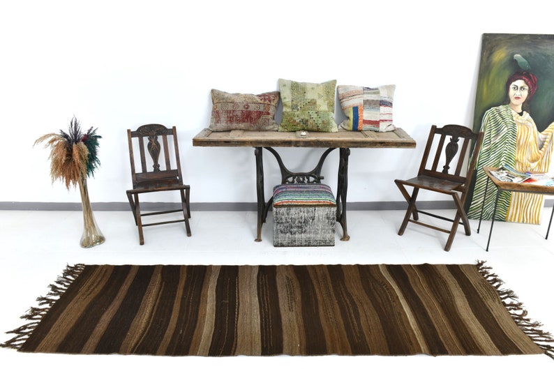May include: A brown striped rug with fringe on the ends. The rug is in front of a wooden table with three pillows on it. There are two wooden chairs and a small stool with a colorful striped cushion on it. A painting of a woman in a yellow and green dress is on the wall.