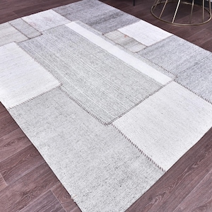 Pode incluir: Um tapete retangular de patchwork em tons de cinza e branco. O tapete é composto por várias peças retangulares e quadradas, costuradas com fio cinza escuro. O tapete é colocado sobre um chão de madeira escura.