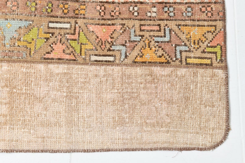 May include: Close-up of the back of a rug with a brown fringe. The rug has a beige background with a colorful geometric pattern.