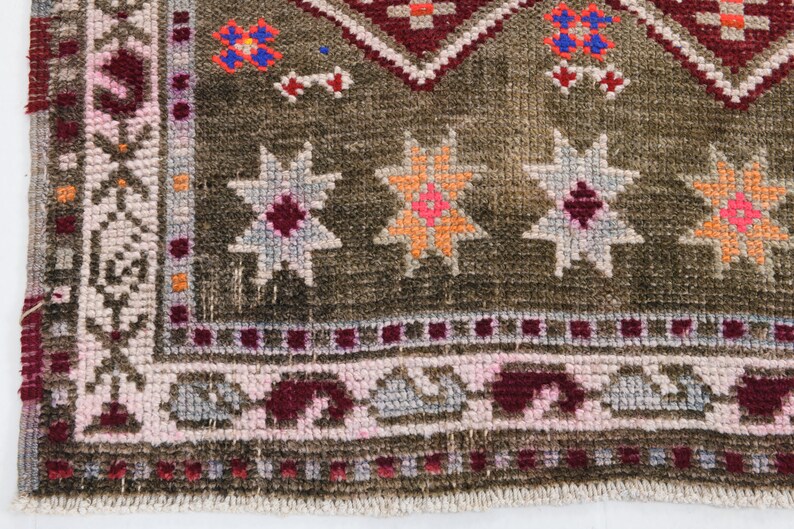 K&ouml;nnte beinhalten: Ein brauner Teppich mit einem geometrischen Muster in Rosa-, Rot-, Orange- und Wei&szlig;t&ouml;nen. Der Teppich hat einen Fransenrand.