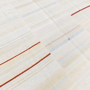 May include: Cream-coloured patchwork rug with a grid pattern. The rug features rectangular patches with subtle variations in tone. Thin, red stripes accent the design, adding a pop of colour to the neutral palette. The rug is made of a woven material.
