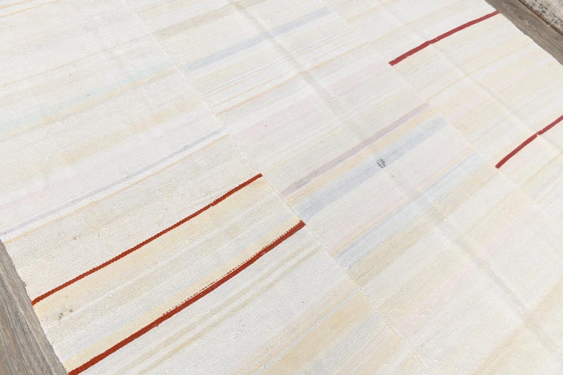 May include: Cream-coloured patchwork rug with a grid pattern. The rug features rectangular patches with subtle variations in tone. Thin, red stripes accent the design, adding a pop of colour to the neutral palette. The rug is made of a woven material.
