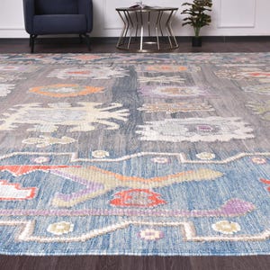 May include: A large, rectangular rug with a blue and grey background. The rug features a repeating pattern of floral and geometric shapes in orange, purple, and cream colours. The rug is made of a woven material and has a textured appearance.