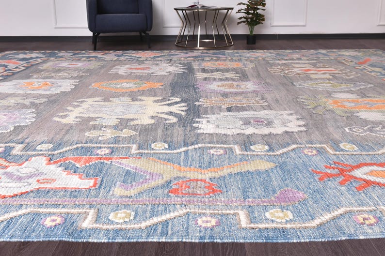 May include: A large, rectangular rug with a blue and grey background. The rug features a repeating pattern of floral and geometric shapes in orange, purple, and cream colours. The rug is made of a woven material and has a textured appearance.