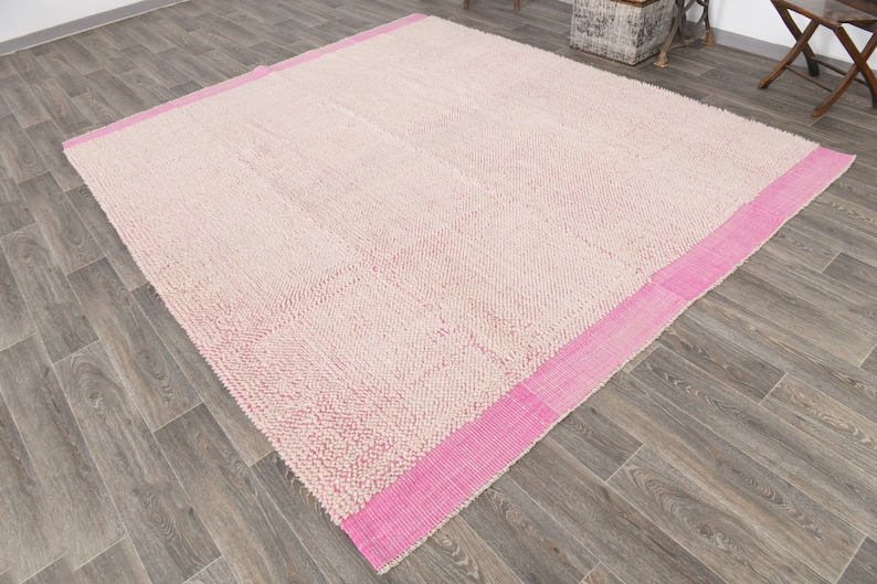 May include: A cream-colored rug with a pink border. The rug has a textured surface and is made of a soft material.
