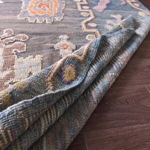 May include: A rolled-up, patterned rug with a grey background and colourful geometric designs in orange, blue, and beige. The rug's edge reveals a blue and beige woven underside, showcasing its texture and construction. The rug is placed on a dark brown wooden floor.