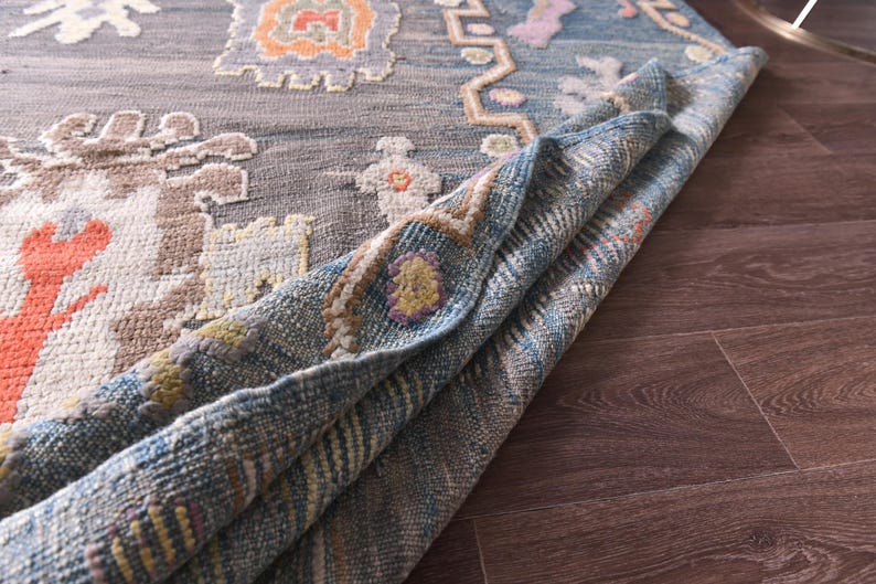 May include: A rolled-up, patterned rug with a grey background and colourful geometric designs in orange, blue, and beige. The rug's edge reveals a blue and beige woven underside, showcasing its texture and construction. The rug is placed on a dark brown wooden floor.
