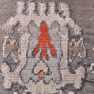 May include: Close-up of a woven textile with a repeating pattern. The design features a central orange figure, surrounded by light blue, grey, and beige geometric shapes. The background is a textured grey.