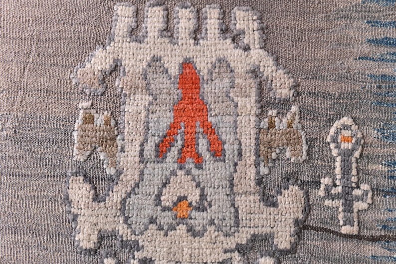May include: Close-up of a woven textile with a repeating pattern. The design features a central orange figure, surrounded by light blue, grey, and beige geometric shapes. The background is a textured grey.
