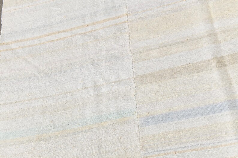 May include: Close-up of a textured, off-white rug with subtle horizontal stripes in beige and light blue. The rug has a soft, neutral colour palette, suitable for various interior styles. The rug's design is simple and elegant.