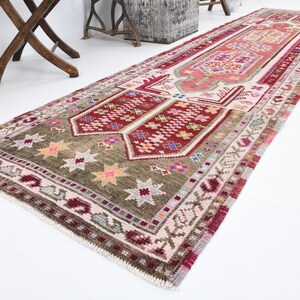 K&ouml;nnte beinhalten: Ein langer, schmaler Teppich mit traditionellem t&uuml;rkischem Design. Der Teppich hat ein rotes, rosa und gr&uuml;nes Farbschema mit komplizierten Mustern und Sternen. Der Teppich ist aus Wolle und hat einen Fransenrand.