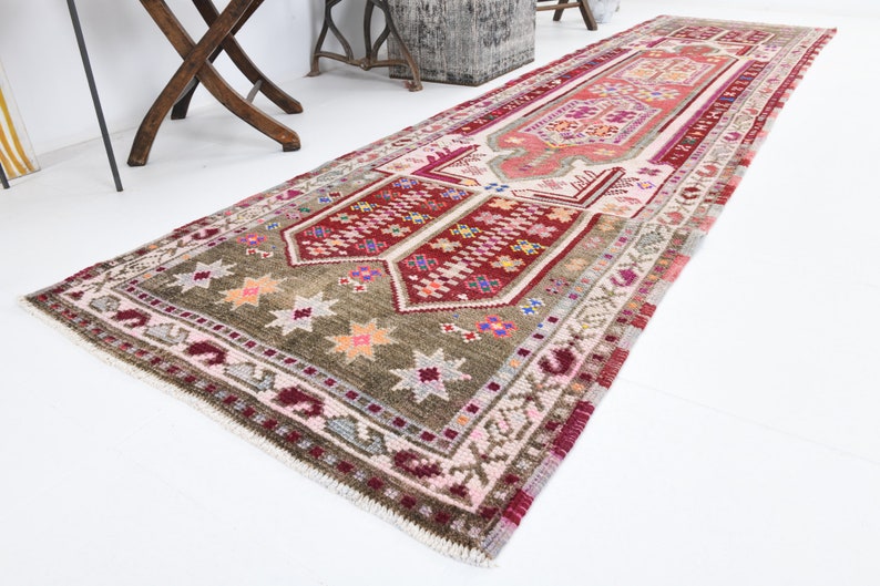 K&ouml;nnte beinhalten: Ein langer, schmaler Teppich mit traditionellem t&uuml;rkischem Design. Der Teppich hat ein rotes, rosa und gr&uuml;nes Farbschema mit komplizierten Mustern und Sternen. Der Teppich ist aus Wolle und hat einen Fransenrand.