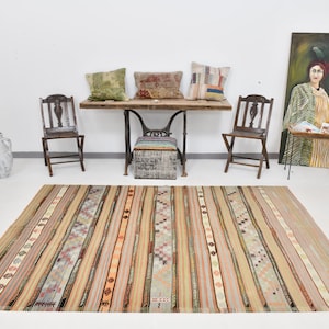 May include: A striped kilim rug with a variety of colors, including beige, brown, orange, and green. The rug is laid out on a white floor in front of a wooden table with three pillows and two chairs.