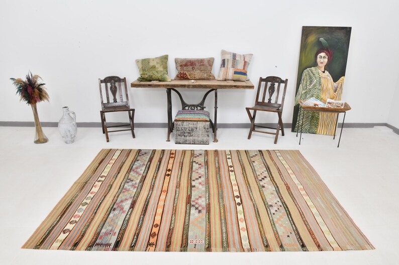 May include: A striped kilim rug with a variety of colors, including beige, brown, orange, and green. The rug is laid out on a white floor in front of a wooden table with three pillows and two chairs.