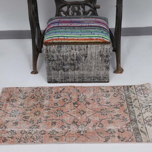 May include: A pink and cream floral patterned rug with a faded look. The rug has a border with a more intricate pattern. The rug is placed on a white tiled floor in front of a black and gray footstool.