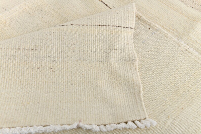 May include: A close-up of a white woven rug with a fringe border. The rug is made of natural fibers and has a subtle texture.