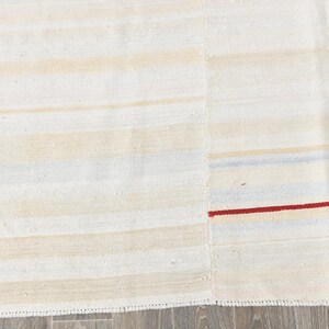 May include: Cream-coloured rug with horizontal stripes in shades of beige and light blue. A thin red stripe accents the design. The rug has a textured appearance and a simple, minimalist style.