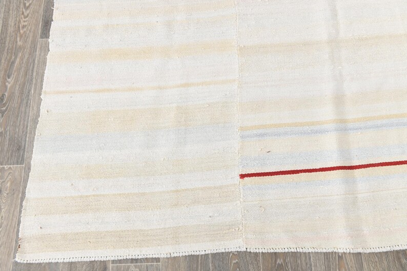 May include: Cream-coloured rug with horizontal stripes in shades of beige and light blue. A thin red stripe accents the design. The rug has a textured appearance and a simple, minimalist style.