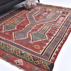 May include: A rectangular, hand-woven rug with a geometric pattern. The rug features a predominantly red background with a central design in shades of grey, green, and blue. The border incorporates yellow, black, and white accents, and the rug has a fringed edge.