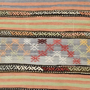 May include: A close-up of a woven rug with a repeating pattern of geometric shapes in shades of red, blue, green, and brown. The rug has a striped design with a central band of intricate patterns.