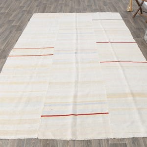 May include: A large, rectangular, off-white rug with horizontal stripes and patches. Thin red lines are evenly spaced across the rug. The rug is made of a woven material and is placed on a wooden floor.