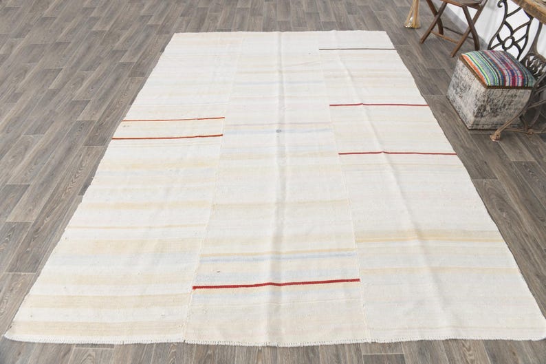 May include: A large, rectangular, off-white rug with horizontal stripes and patches. Thin red lines are evenly spaced across the rug. The rug is made of a woven material and is placed on a wooden floor.