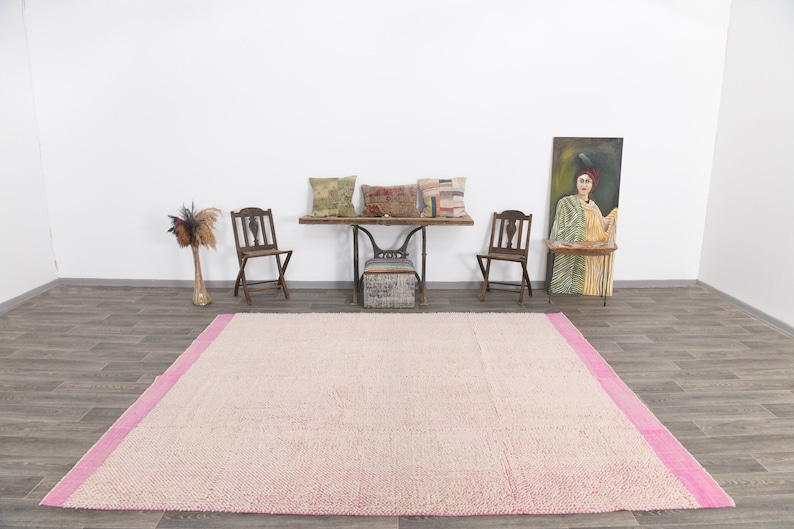 May include: A light pink and white area rug with a pink border. The rug is on a wooden floor in a room with two chairs, a table, and a painting.