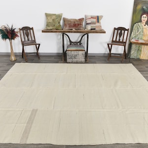 May include: A large, off-white, rectangular rug with a subtle woven pattern. The rug is laid out on a wood floor in a room with a wooden table and chairs.