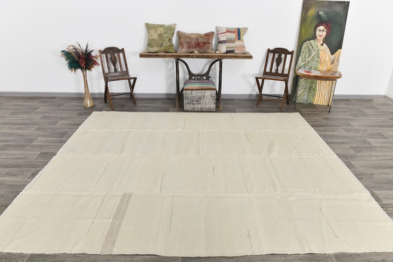 May include: A large, off-white, rectangular rug with a subtle woven pattern. The rug is laid out on a wood floor in a room with a wooden table and chairs.