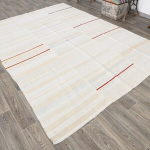 May include: A large, rectangular, off-white rug with a patchwork design. The rug features thin, red stripes and subtle blue accents. The rug is placed on a wooden floor.