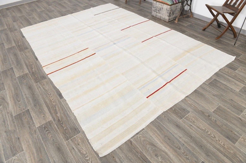 May include: A large, rectangular, off-white rug with a patchwork design. The rug features thin, red stripes and subtle blue accents. The rug is placed on a wooden floor.