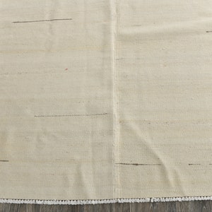 May include: A white woven rug with a subtle texture and a fringe border. The rug has a few thin black lines woven into the design.