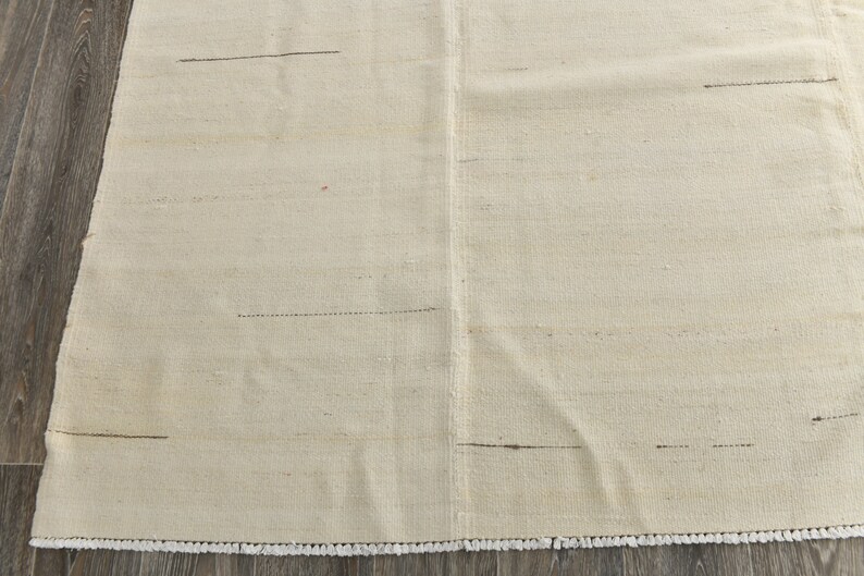 May include: A white woven rug with a subtle texture and a fringe border. The rug has a few thin black lines woven into the design.