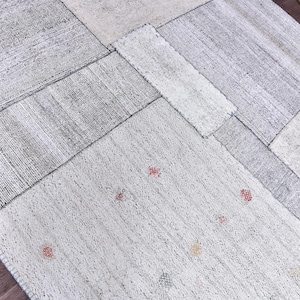 Pode incluir: Um tapete patchwork com uma paleta de cores neutras. O tapete apresenta vários retalhos retangulares e quadrados em tons de cinza e branco sujo. Pequenos detalhes em vermelho e amarelo estão espalhados pela superfície, adicionando um toque de cor.