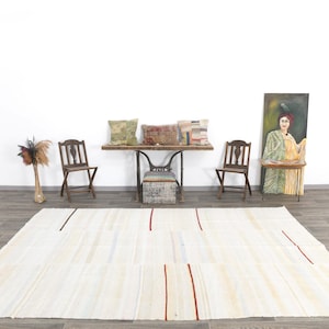 May include: A large, rectangular, cream-coloured rug with vertical red and black stripes. The rug is placed on a wooden floor in a room with a white wall. The room also features wooden chairs, a table, and decorative cushions.