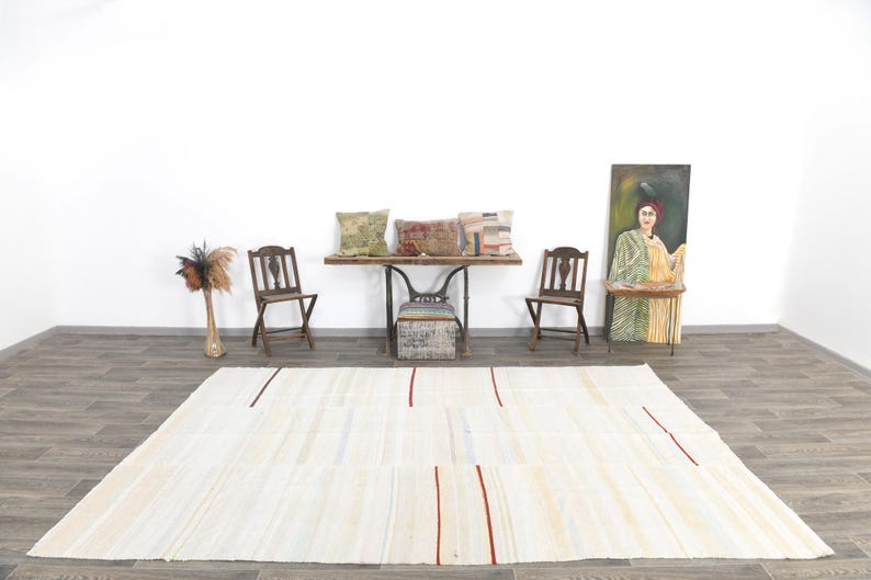 May include: A large, rectangular, cream-coloured rug with vertical red and black stripes. The rug is placed on a wooden floor in a room with a white wall. The room also features wooden chairs, a table, and decorative cushions.