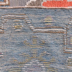 May include: Close-up of a section of a woven rug with a blue background and geometric patterns in brown, yellow, and purple. The rug has a textured appearance, with visible threads and a detailed design. The rug's edge is visible.